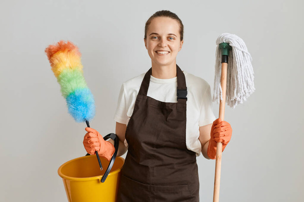 Indoor shot of Caucasian housewife cleaning disinfecting all surfaces, holding mop, bucket and pp duster, cleaning house, wearing gloves, apron, t-shirt and jeans. - Photo, Image