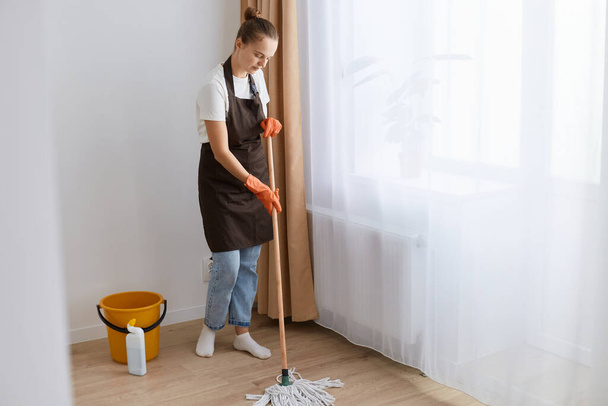 Indoor shot of dark haired female with hair bun wearing white t shirt, apron and jeans cleaning her flat, washing floor with mop, doing household chores. - Fotoğraf, Görsel