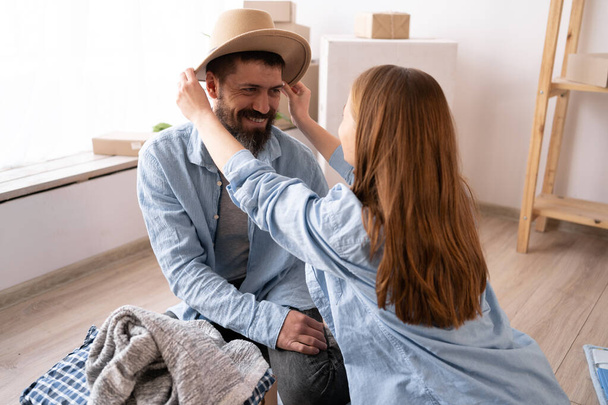 a young cheerful couple in a new apartment on the day of the move sit on the floor unpack things the woman puts on a hat to the man. moving to a new apartment. Lots of packing boxes around them. - Foto, Imagen