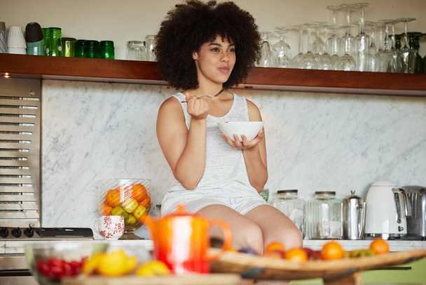 I bathe in nothing but pride. Shot of a beautiful young woman sitting on a countertop and having breakfast in the kitchen at home. - Фото, изображение