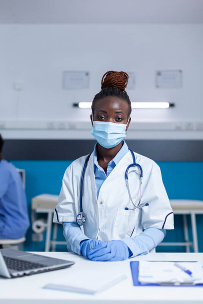 Νοσοκομειακός ιατρός που κάθεται στο τραπέζι του γραφείου φορώντας μάσκα προστασίας από ιούς και χειρουργικά γάντια - Φωτογραφία, εικόνα