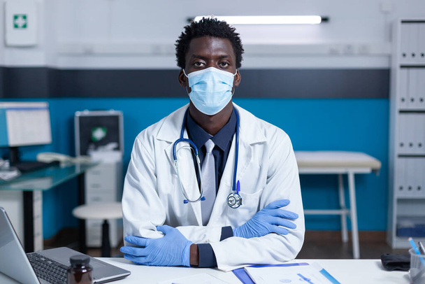 African american hospital doctor sitting at office desk while wearing facemask because of coronavirus pandemic - Valokuva, kuva