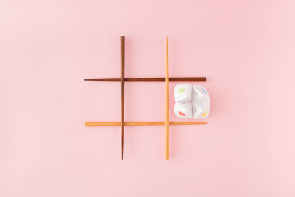 Tic tac toe game with wooden food chopsticks and paper origami fortune teller on pink background. Minimal concept. - Фото, изображение