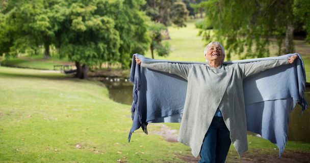 Cheerful caucasian senior woman holding scarf with arms outstretched against trees in park. nature, happy, unaltered, senior citizens, retirement and awareness concept. - 写真・画像