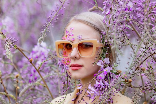 Beautiful woman in sunglasses and with flowers petals on face around blooming wisteria tree branches - Photo, Image