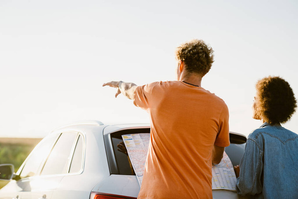 Young multiracial couple examining map and pointing hand away during car trip - Fotó, kép