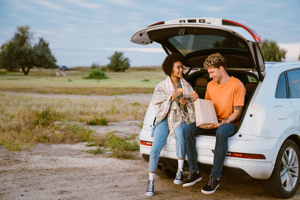 Young multiracial couple eating peach while sitting in trunk during car trip - Фото, зображення