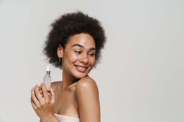 Black curly brunette woman smiling while posing with face serum isolated over white background - Фото, изображение