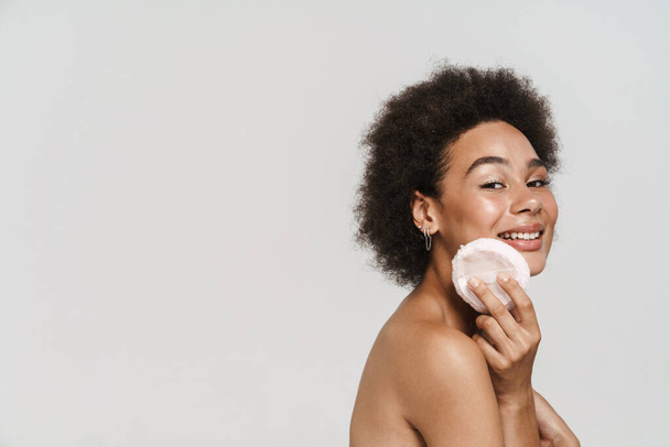 Black curly shirtless woman smiling while using powder sponge isolated over white background - Photo, image
