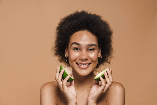 Black curly shirtless woman smiling while posing with avocado isolated over beige background - 写真・画像