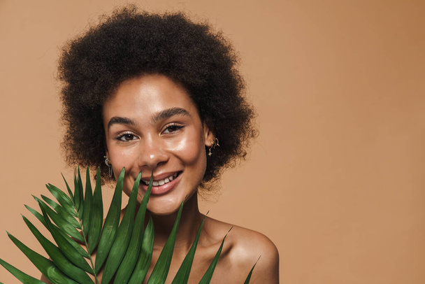 Black shirtless woman smiling while posing with green leaf isolated over beige background - Foto, Imagen