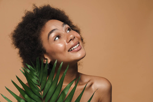 Black shirtless woman smiling while posing with green leaf isolated over beige background - Foto, Bild