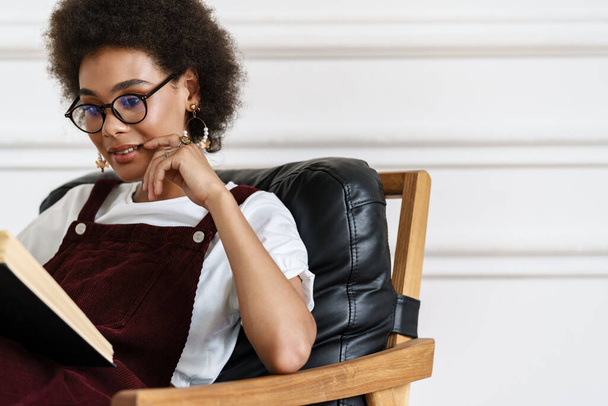Young black woman wearing eyeglasses reading book while sitting on armchair at home - Foto, immagini