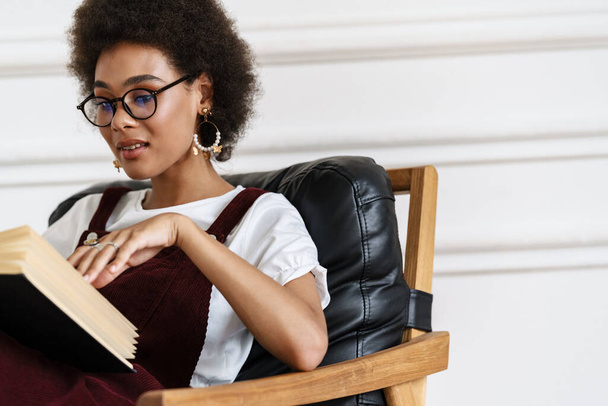 Young black woman wearing eyeglasses reading book while sitting on armchair at home - Фото, изображение