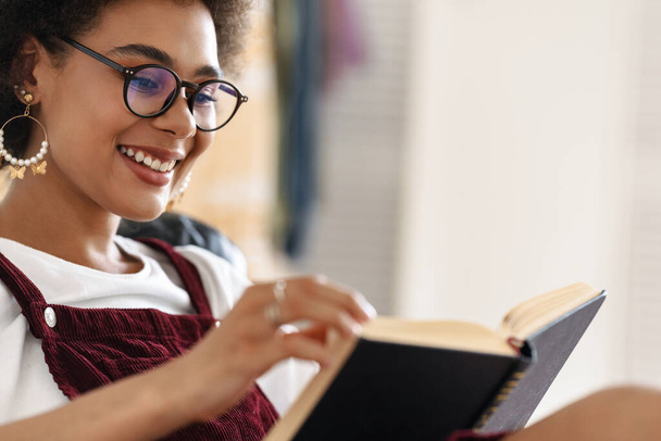 Young black woman wearing eyeglasses smiling and reading book while resting at home - Photo, image