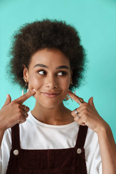 Young black woman smiling and pointing fingers at her cheeks isolated over blue background - Foto, Bild