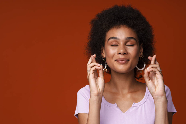 Young black woman smiling while posing with fingers crossed isolated over brown background - Photo, image