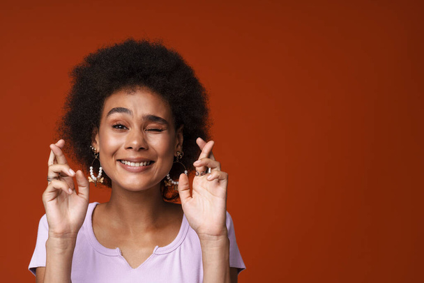 Young black woman winking while posing with fingers crossed isolated over brown background - Photo, image