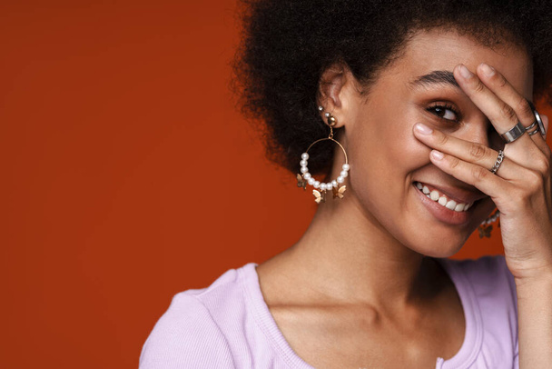 Young black woman wearing earrings smiling and covering her face isolated over brown background - Fotó, kép
