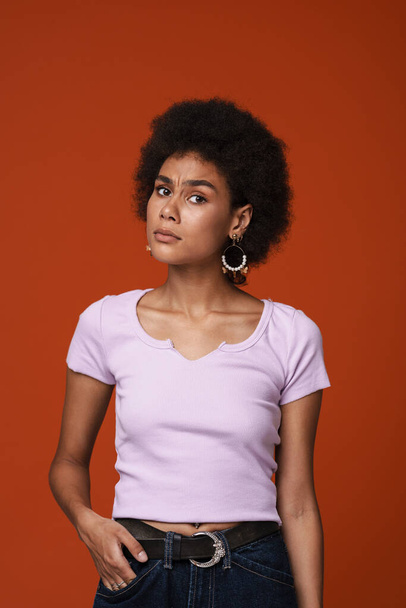 Black young woman in earrings frowning and looking at camera isolated over brown background - Foto, Imagem
