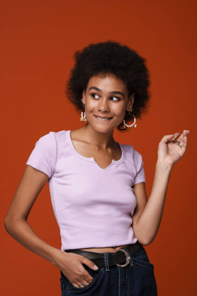 Black young woman in earrings gesturing and smiling at camera isolated over brown background - Fotó, kép