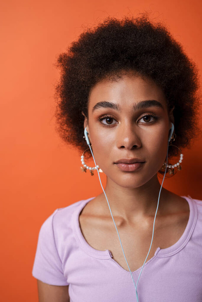Black young woman in earrings listening music with earphones isolated over brown background - Foto, afbeelding