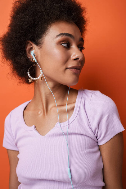 Black young woman in earrings listening music with earphones isolated over brown background - Foto, Imagem