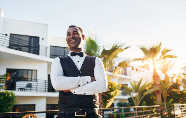 Posing for a camera. Black waiter in formal clothes is at his work outdoors at sunny daytime. - Photo, image