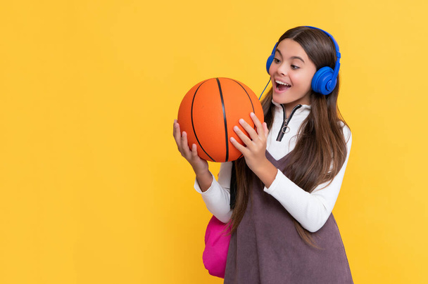 zszokowany dzieciak w słuchawkach ze szkolnym plecakiem i piłką do koszykówki na żółtym tle - Zdjęcie, obraz