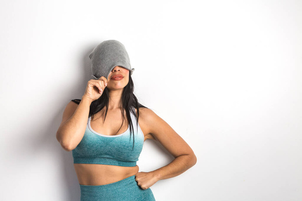 Fit female model dressed in sports bra and leggings covering half of face with beanie while standing with hand on waist on white background - Photo, Image