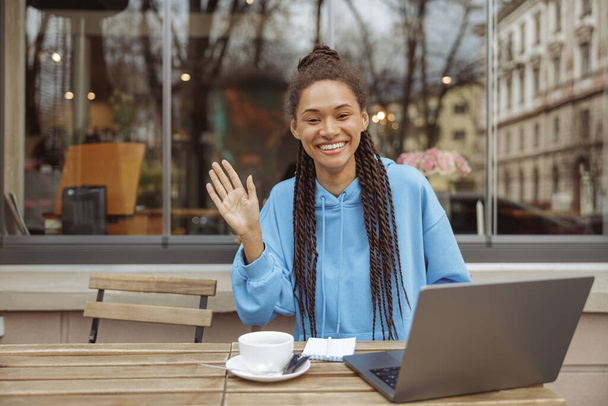 Portret van jonge vrolijke gemengde ras stijlvolle vrouwelijke freelancer zwaaien met de hand op laptop. - Foto, afbeelding