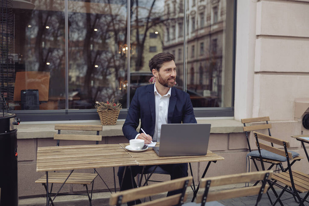 Yakışıklı bir iş adamı, açık kafede dizüstü bilgisayar başında çalışırken not alıyor. Kahve molası. - Fotoğraf, Görsel