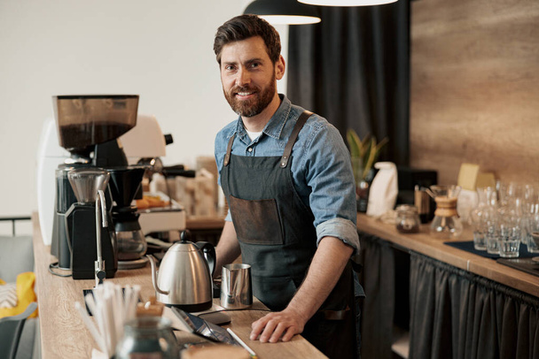 Barista con barba elegante sonriendo mientras está detrás de un mostrador en el coffeeshop - Foto, imagen