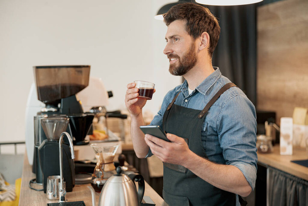 barista wearing apron drinking coffee and holding phone during lunch break - Foto, Bild