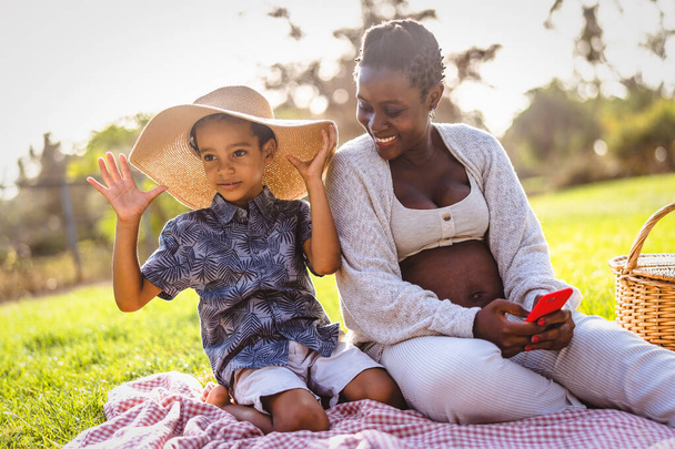 Ευτυχισμένη αφρικανή έγκυος μητέρα περνάει χρόνο με το γιο του κάνοντας πικ-νικ κατά τη διάρκεια του Σαββατοκύριακου στο δημόσιο πάρκο - Η μητρότητα και οι γονείς έννοια του τρόπου ζωής - Φωτογραφία, εικόνα