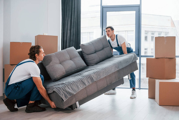 Carrying heavy sofa. Two young movers in blue uniform working indoors in the room. - Fotoğraf, Görsel