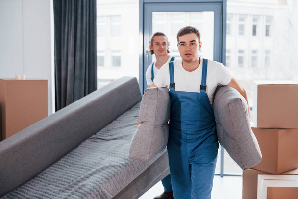 Carrying heavy sofa. Two young movers in blue uniform working indoors in the room. - Photo, Image