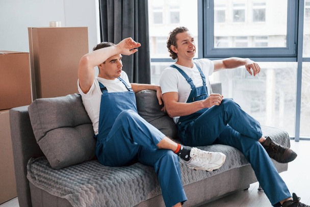 Sitting on the sofa. Tired and taking break. Two young movers in blue uniform working indoors in the room. - Foto, imagen