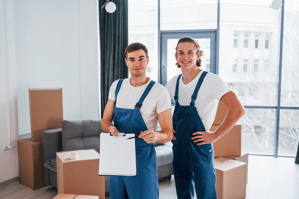 Holds notepad with document. Two young movers in blue uniform working indoors in the room. - Foto, Imagen