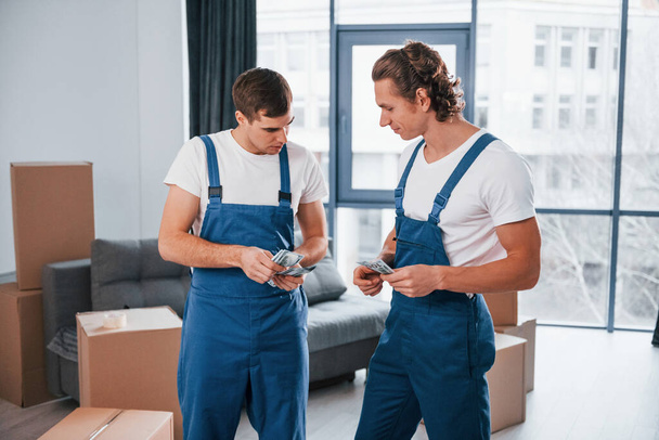 Holding money. Two young movers in blue uniform working indoors in the room. - Foto, Bild
