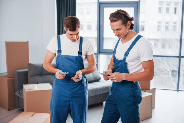 Holding money. Two young movers in blue uniform working indoors in the room. - 写真・画像