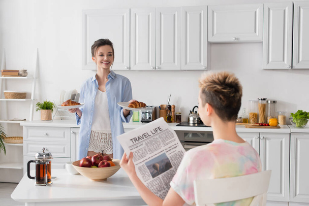cheerful pansexual person holding croissants near partner with travel life newspaper - Valokuva, kuva