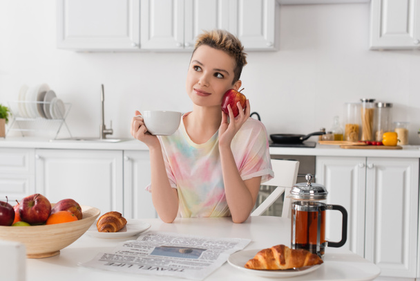 smiling pansexual person with apple and cup of tea sitting near croissants and newspaper - Foto, Imagem