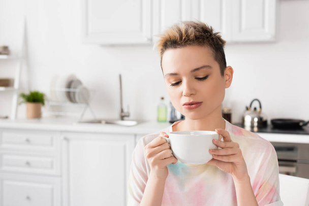 young bigender person with short hair holding cup of tea in blurred kitchen - Fotografie, Obrázek