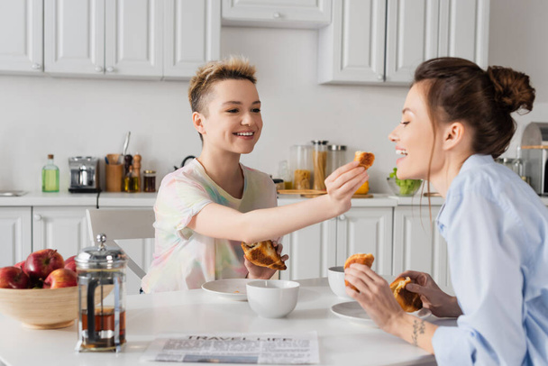 smiling bigender person feeding partner with croissant during breakfast in kitchen - Photo, Image