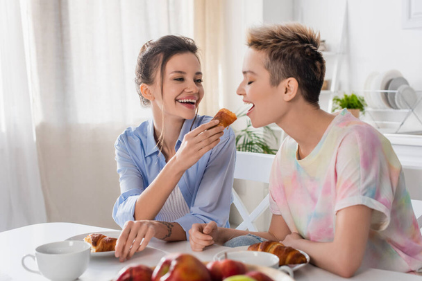 cheerful pangender person feeding lover with croissant while having breakfast in kitchen - Foto, afbeelding