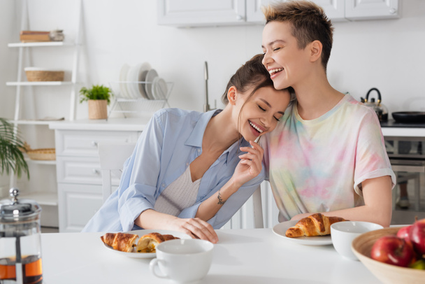 excited pansexual partners laughing in kitchen near croissants and tea cups - Photo, image