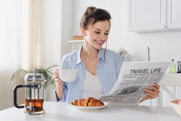 smiling woman with cup of tea reading travel life newspaper near delicious croissant - Photo, image