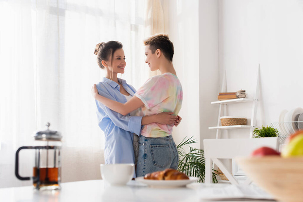 happy bigender couple embracing in kitchen near blurred breakfast - 写真・画像