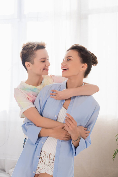 joyful bigender people embracing and smiling at each other at home - Фото, изображение